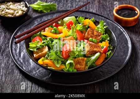 Tofu-Salat mit knackigen Salatblättern, grünen Erbsen, Tomaten, gelbem Paprika in schwarzer Schüssel mit Essstäbchen auf dunklem Holztisch, Nahaufnahme Stockfoto