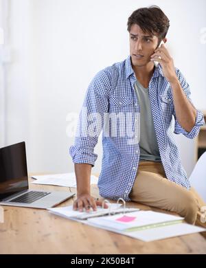 Er verwendet alle ihr zur Verfügung stehenden Werkzeuge. Hübscher junger ethnischer Geschäftsmann, der mit seinem Laptop und Papierkram vor ihm telefoniert. Stockfoto