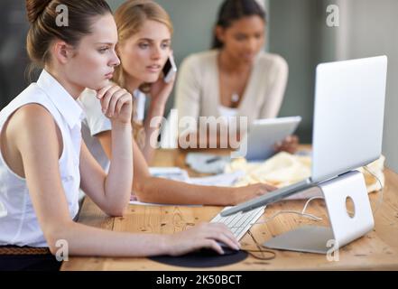 Entscheidung für den Look für die nächste Saison. Drei junge Modedesigner, die an einem Laptop arbeiten und sich auf einem Handy unterhalten. Stockfoto