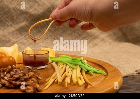 Verschiedene Käsesorten, Walnusskerne, Honig in einer Glasvase und geräucherte Wachteleier auf einem Holzbrett. Jemand taucht ein Stück Käse in Honig. Stockfoto