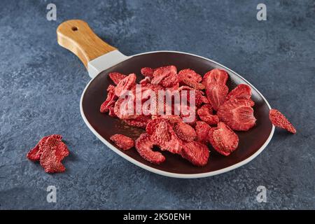 Erdbeersplitter werden in Keramikschale mit Holzgriff auf grau-blauem Betongrund gestapelt. Hundertprozentig natürliche gefriergetrocknete Erdbeeren Stockfoto