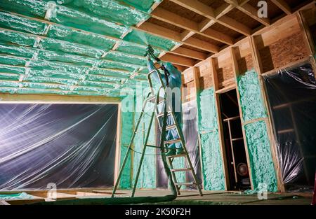 Männlich Baumeister isolierenden Holzrahmen Haus. Mann Arbeiter sprühen Polyurethan-Schaum in der zukünftigen Hütte, stehend auf der Leiter, mit Plural Komponente Pistole. Bau- und Isolierkonzept. Stockfoto