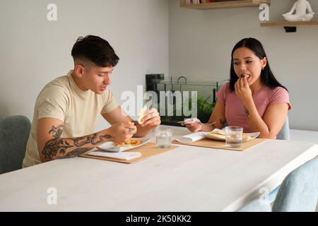 Zwei junge lateinische Menschen essen traditionelle Arepas im Wohnzimmer. Stockfoto