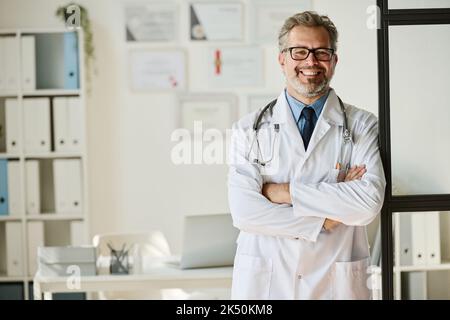 Porträt eines professionellen medizinischen Spezialisten im weißen Mantel, der mit gekreuzten Armen steht und die Kamera im Büro anlächelt Stockfoto
