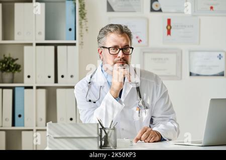 Porträt eines reifen Arztes in weißem Mantel, der die Kamera anschaut, während er mit einem Laptop im Büro an seinem Arbeitsplatz sitzt Stockfoto
