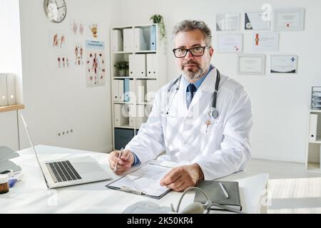 Porträt eines reifen Arztes in einer Brille, die medizinische Dokumente am Tisch im Büro ausfüllt Stockfoto
