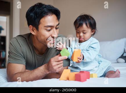 Baby, Down-Syndrom und Lernen auf einem Bett mit Kind und Vater spielen mit pädagogischen Blöcken in einem Schlafzimmer. Familie, Behinderung und Kinder, die sich mit uns verbinden Stockfoto