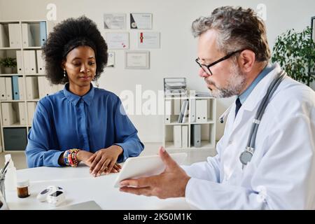 Die reife Ärztin zeigt den Patienten die Analyseergebnisse auf einem digitalen Tablet während ihrer Konsultation im Büro Stockfoto