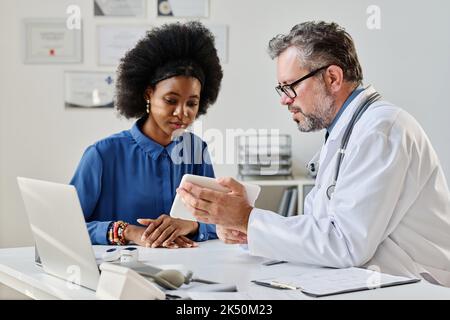 Reifer Arzt, der einer Patientin während ihres Krankenhausaufenthalts die Diagnose online auf einem digitalen Tablet zeigt Stockfoto