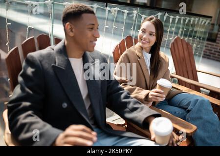 Lächelndes interrassisches Paar in Mänteln, das mit Pappbechern auf Holzsitzen im Freien sitzt, Stockbild Stockfoto