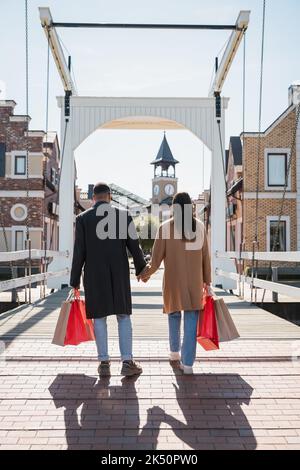 Rückansicht eines multiethnischen Paares in Mänteln, das mit Einkaufstaschen über die Brücke läuft und die Hände hält, Stockbild Stockfoto