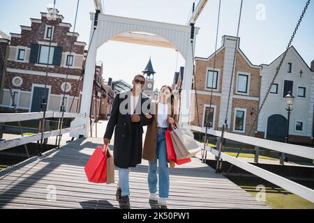 Fröhliches, multiethnisches Paar in stilvoller Kleidung und Sonnenbrille, das mit Einkaufstaschen über die Brücke läuft, Stockbild Stockfoto