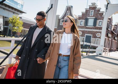 Fröhliches und modisches Paar in Sonnenbrillen, das die Hände hält, während es mit Einkaufstaschen über die Stadtbrücke läuft, Stockbild Stockfoto