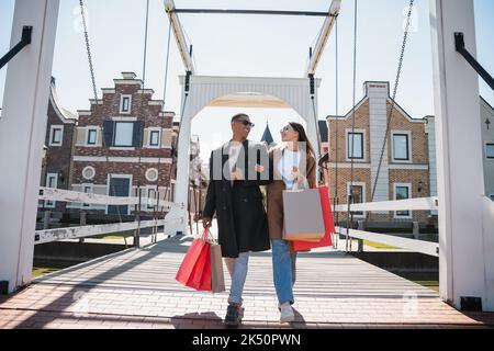 Stilvolles interracial Paar lächelt sich an und läuft mit Einkaufstaschen über die Brücke, Stockbild Stockfoto