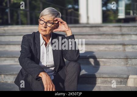 Eine Geschäftsfrau, die vor dem Firmengebäude sitzt. Sie ist nachdenklich. Stockfoto