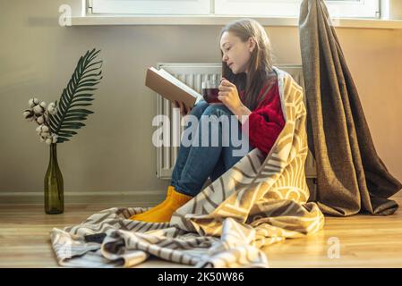 Eine Frau, in ein kariert gehüllt, sitzt mit heißem Tee auf dem Boden und wärmt sich neben einem warmen Heizkörper auf. Das Konzept der Wirtschaft und die Krise der ENE Stockfoto