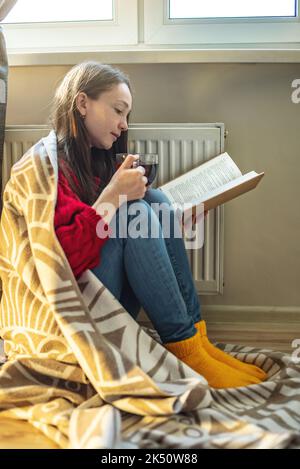 Eine Frau, in ein kariert gehüllt, sitzt mit heißem Tee auf dem Boden und wärmt sich neben einem warmen Heizkörper auf. Das Konzept der Wirtschaft und die Krise der ENE Stockfoto