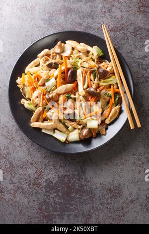 Hacken Sie das gebratene Huhn mit Pilzen, Bok Choy, Zwiebeln, Bohnensprossen und Karotten in Nahaufnahme auf einem Teller auf dem Tisch. Vertikale Draufsicht von oben Stockfoto