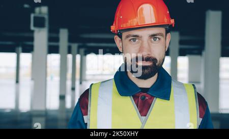 Porträt eines Baumeisters in Uniform, der die Kamera ansah und lächelnd auf den Kopf nickte, der im Gebäude stand. Mitarbeiter- und Arbeitskonzept. Stockfoto
