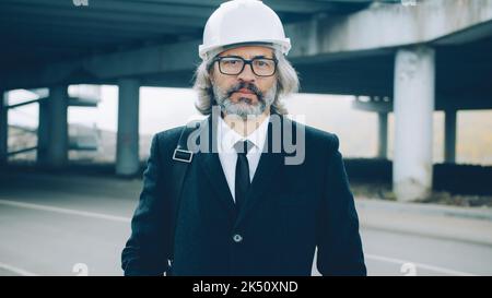 Porträt eines respektvollen Geschäftsmannes, der im Freien in der Industriezone steht und Schutzhelm und eleganten Anzug trägt und die Kamera mit ernstem Gesicht anschaut Stockfoto