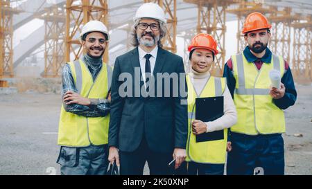 Verschiedene Gruppen von Menschen, die ein Team bauen, stehen auf der Baustelle und blicken lächelnd auf die Kamera, die Helme und Sicherheitswesten trägt Stockfoto
