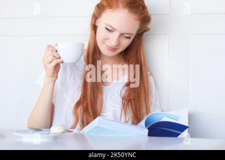 Sie genießt es, sich mit einem guten Buch zu entspannen. Eine attraktive junge Frau, die ein Buch liest und Kaffee trinkt. Stockfoto