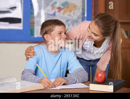 Ich nehme mir Zeit für alle meine Schüler. Eine unterstützende junge Lehrerin, die sich über ihren Schüler lehnt, während er in einem Buch schreibt. Stockfoto