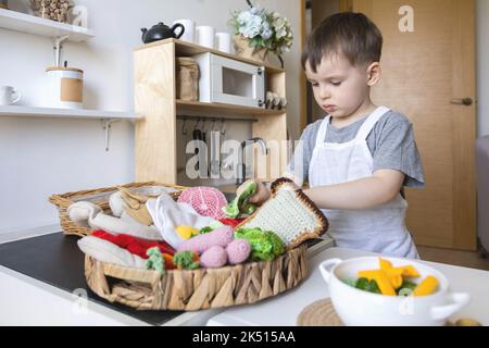 Niedlicher kleiner Junge mit Kochmütze und Schürze, der in der kindischen Küche beim Kochen von Speisen spielt Stockfoto