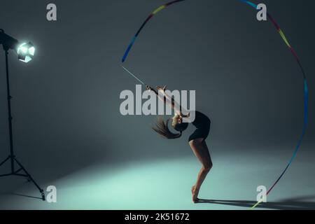 Turnerin Kind Mädchen in schwarzem Bodysuit führt Gymnastik-Übungen mit Band in der Hand durch Scheinwerfer auf dunklem Hintergrund beleuchtet. Stockfoto