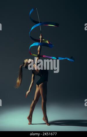 Turnerin Kind Mädchen von Licht in schwarzem Bodysuit beleuchtet führt Gymnastik-Übungen mit Band in der Hand auf dunklem Hintergrund. Stockfoto