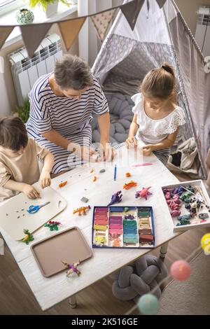 Glückliche junge Großmutter Formen Dinosaurier aus Plastilin spielen Enkel und Enkelin genießen Familienwochenende Stockfoto