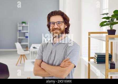 Selbstbewusster kaukasischer Mann mit lächelndem Bart und Blick auf die Kamera, die im Büro mit gekreuzten Armen posiert Stockfoto