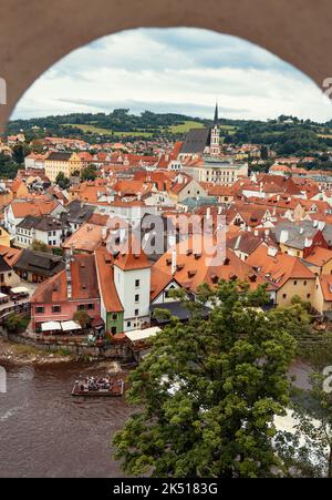 Der Blick auf die St. Vitus Kirche und die Stadt Cesky Krumlov vom Schloss aus durch das Fenster in der Wand. Menschen auf Holzfloß auf der Moldau. Stockfoto