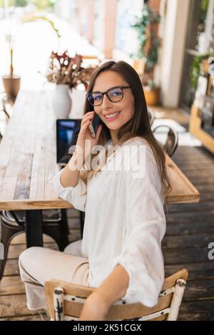 Ein großer Winkel von glücklicher Unternehmerin in leichter Kleidung und Brille, die mit einem Lächeln auf die Kamera schaut und auf einen Anruf antwortet, während sie auf einem Holztisch sitzt Stockfoto