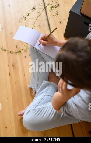 Draufsicht auf eine nicht erkennbare Frau in legerer Kleidung, die Daten im Notebook aufschreibt und auf dem Mobiltelefon spricht, während sie mit gekreuzten Beinen auf Holzboden sitzt Stockfoto