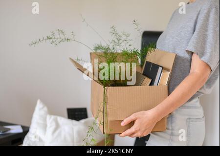 Nicht erkennbare Frau in legerer Kleidung, die Pappkarton mit Büchern und Topfpflanzen trägt, während sie in eine neue Wohnung umzieht Stockfoto