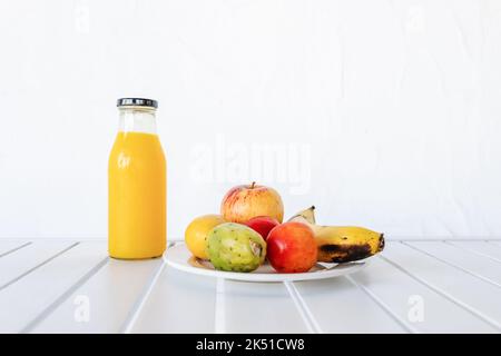 Eine Flasche frischen Orangensaft auf dem weißen Tisch neben dem Teller mit verschiedenen bunten reifen Früchten bei Tageslicht Stockfoto
