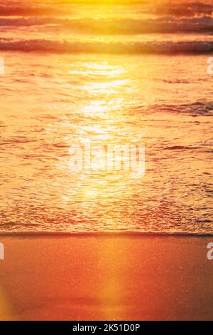 Sonnenuntergang über dem Meer. Riplpe Meer Ozean Wasseroberfläche mit kleinen Wellen. Ozean Wasser Schaum Spritzwasser waschen Sandstrand. Vertikales Foto Stockfoto