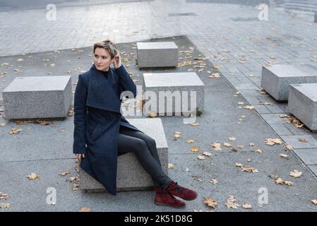 Auf dem Stadtplatz sitzt eine kurzhaarige, attraktive Blondine in einem Mantel und roten Schuhen. Stockfoto