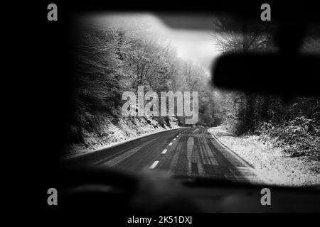 Schwarz-Weiß-Ernte unkenntlich Person, die das Auto entlang der asphaltierten Straße zwischen blattlosen Bäumen fährt, die mit Reif bedeckt sind, am bewölkten Wintertag Stockfoto
