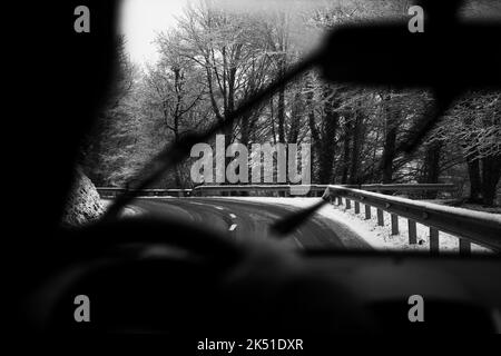 Schwarz-Weiß-Ernte unkenntlich Person, die das Auto entlang der asphaltierten Straße zwischen blattlosen Bäumen fährt, die mit Reif bedeckt sind, am bewölkten Wintertag Stockfoto