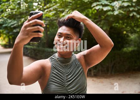 Glückliche philippinische Transgender-Frau lächelt und berührt kurze Haare, während sie am Sommerwochenende im Park Selfie per Smartphone macht Stockfoto