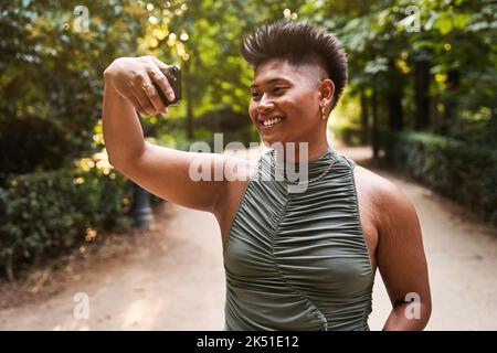 Glückliche philippinische Transgender-Frau lächelt und berührt kurze Haare, während sie am Sommerwochenende im Park Selfie per Smartphone macht Stockfoto