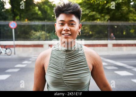 Fröhliche asiatische Transgender Frau mit kurzen Haaren und Pickel im Gesicht Blick auf die Kamera, während sie auf verschwommenem Hintergrund der Straße und Park in c stehen Stockfoto