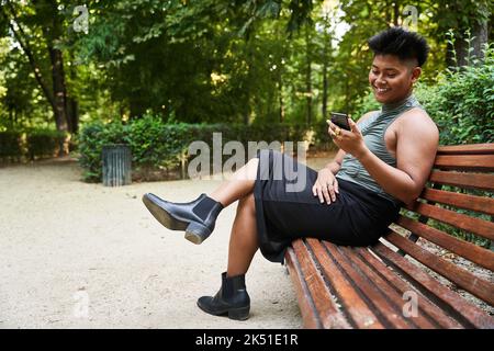 Ganzkörper glücklich asiatische nicht binäre Person in femininer Kleidung lächelnd und beobachten Video auf dem Handy, während auf der Bank im Sommer Park sitzen Stockfoto