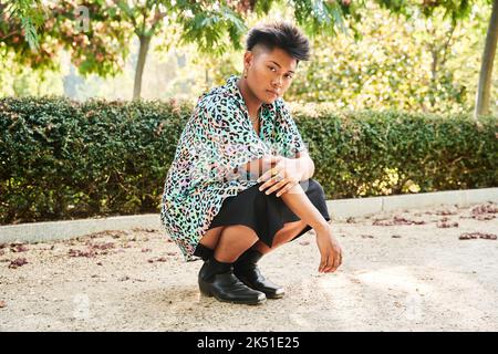 Ganzkörper asiatische nicht binäre Person in stilvollem Hemd und Rock Blick auf die Kamera, während auf Spukken am sonnigen Sommertag im Park sitzen Stockfoto