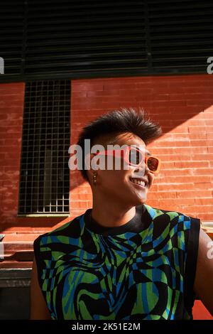 Von unten glücklich philippinischen nicht binären Person in stilvoller Kleidung und Sonnenbrille Blick weg und lächeln gegen sonnenbeschienenen Ziegelwand Stockfoto