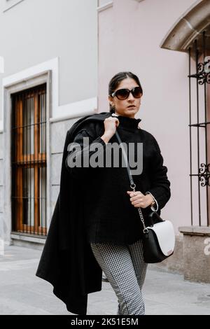 Selbstbewusste junge hispanische Frau mit dunklem Haar in schwarzem Pullover und Sonnenbrille, die mit Mantel über Schulter auf der Stadtstraße läuft Stockfoto