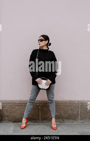 Ganzer Körper von ernst selbstbewussten erwachsenen ethnischen Frauen mit langen dunklen Haaren in stilvollem Outfit hohen Absätzen Schuhe und Sonnenbrille auf der Straße in der Nähe grau stehen Stockfoto