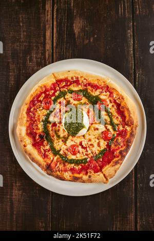 Von oben köstliche Pizza mit Kirschtomaten und Parmesan garniert mit Mozzarella-Käseball und Pesto-Sauce und serviert auf dem Teller auf dem Holztisch Stockfoto